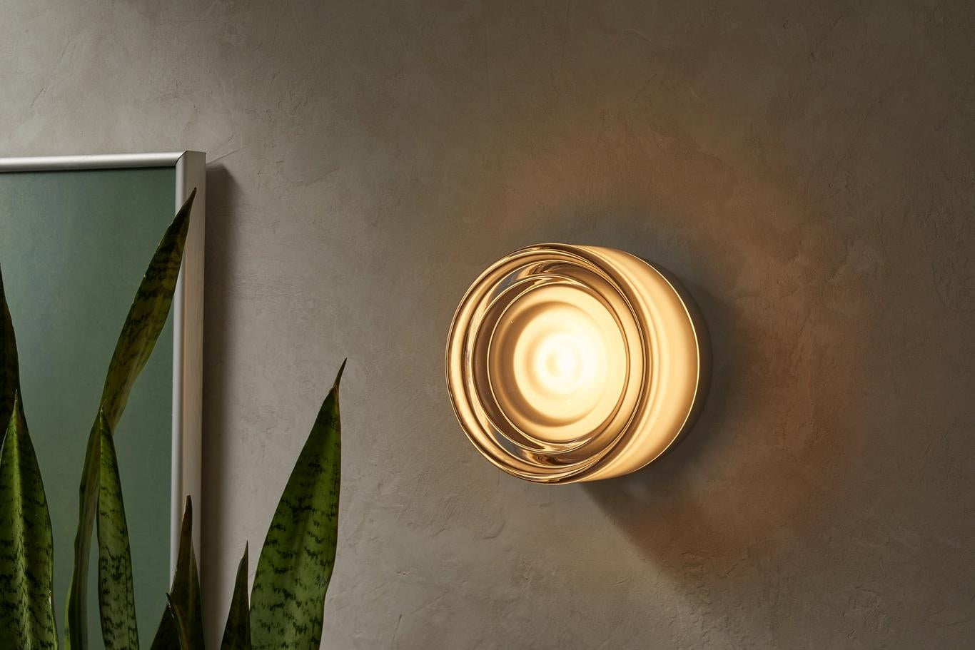 Wall-mounted gold light fixture on a neutral wall with a plant in the corner